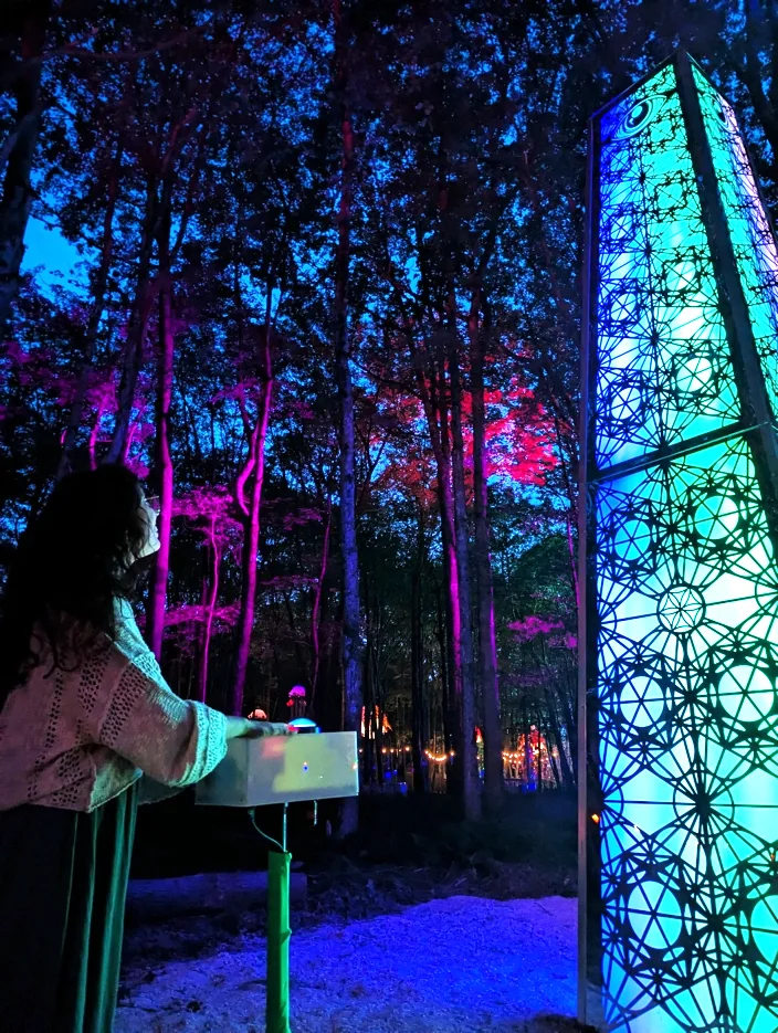 A visitor controlling The Obelisk’s illuminated geometric tower amid trees glowing blue and pink.